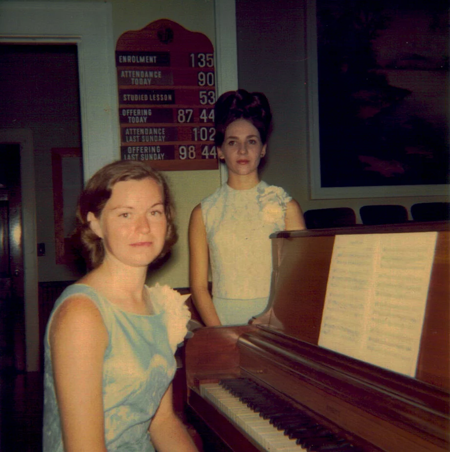Two young women at the church piano beside the Sunday attendance board.